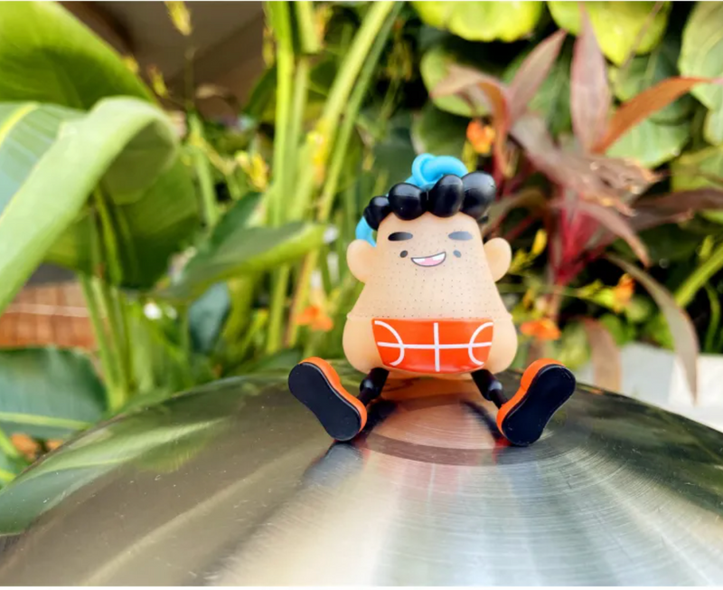 Cute basketball-themed figurine with blue hair, sitting on metal surface amid green plants, perfect family gift