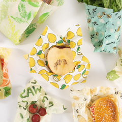 Colorful reusable food wraps with lemon, leaf, and bee prints wrapping apples, tomatoes, and sandwiches