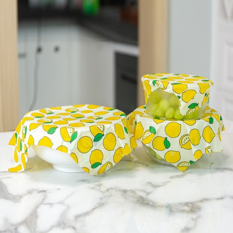 Lemon-patterned reusable food wraps covering bowls on marble countertop, eco-friendly kitchen storage